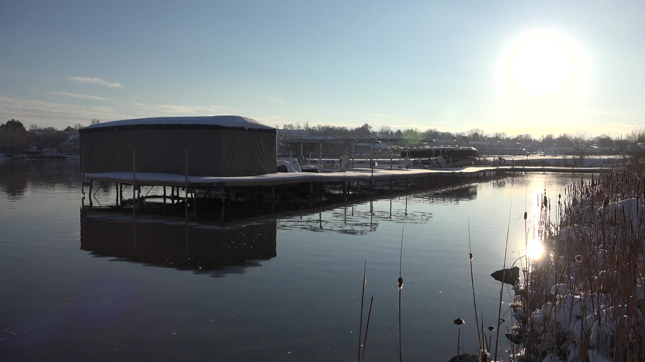 A covered dock sits at the left edge of a Westminster, Colorado lagoon, with reeds at right, snow along the bank, strong sun glare, and no visible people.