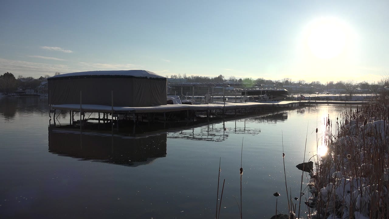 Wide view in Westminster, Colorado of a tan covered structure on a white platform reflected in calm water, with reeds at right and no visible people.