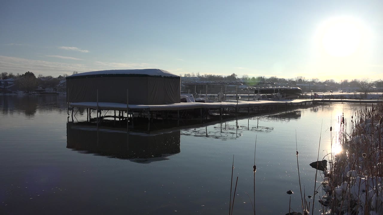 Covered dock on a still pond with bright sun reflection and reeds at right.