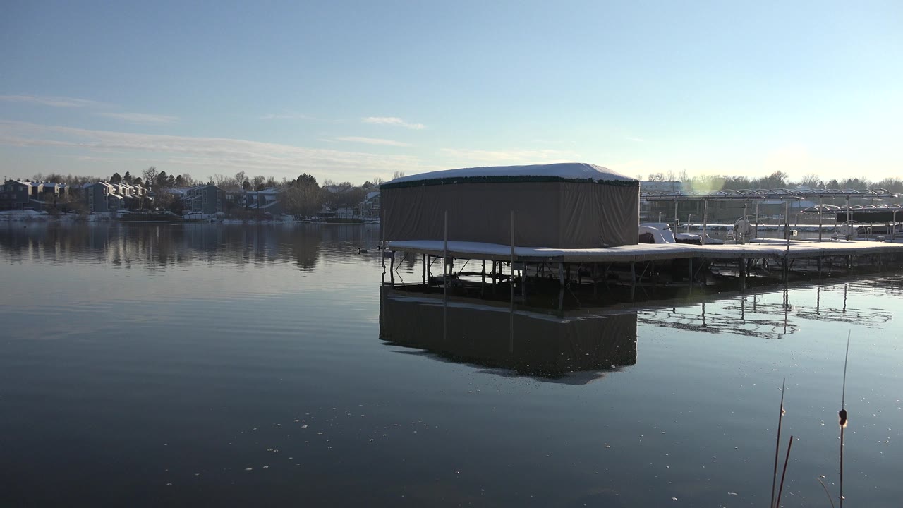 At 0:00 in Westminster, Colorado, a covered dock building sits over calm reflective water with distant houses, bare trees, pale sky, and no visible people.