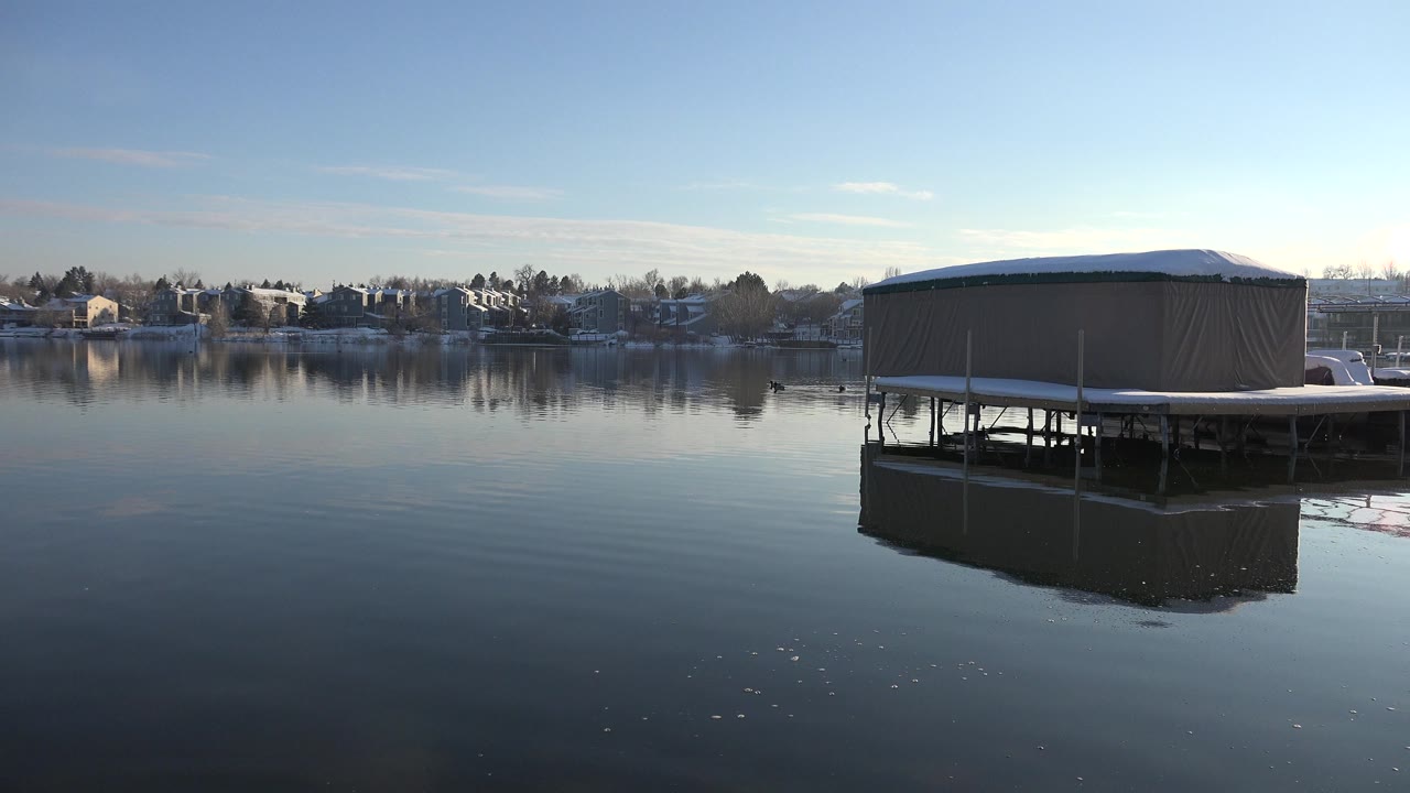 Covered tan platform over calm water with shoreline homes in the distance.