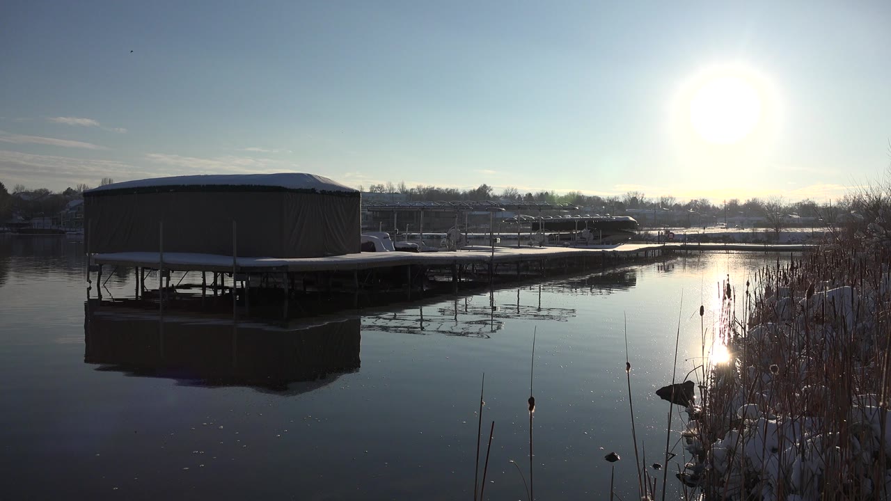 Covered dock or boat lift on still water in Westminster, Colorado, with reeds at the right edge, snow along the shore, bright sun glare, and no visible people.