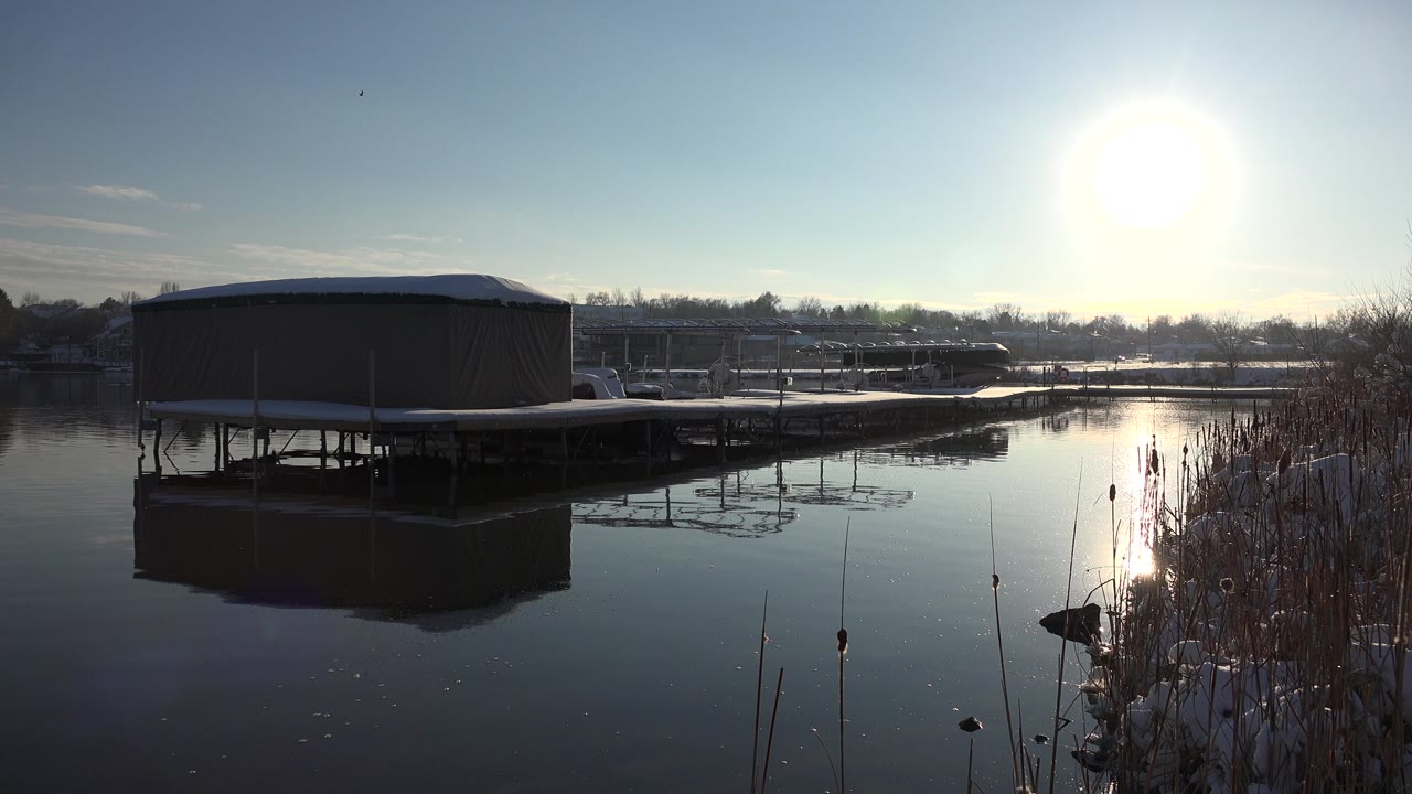 Sunlit water, covered dock, and reeds along a snowy shoreline.