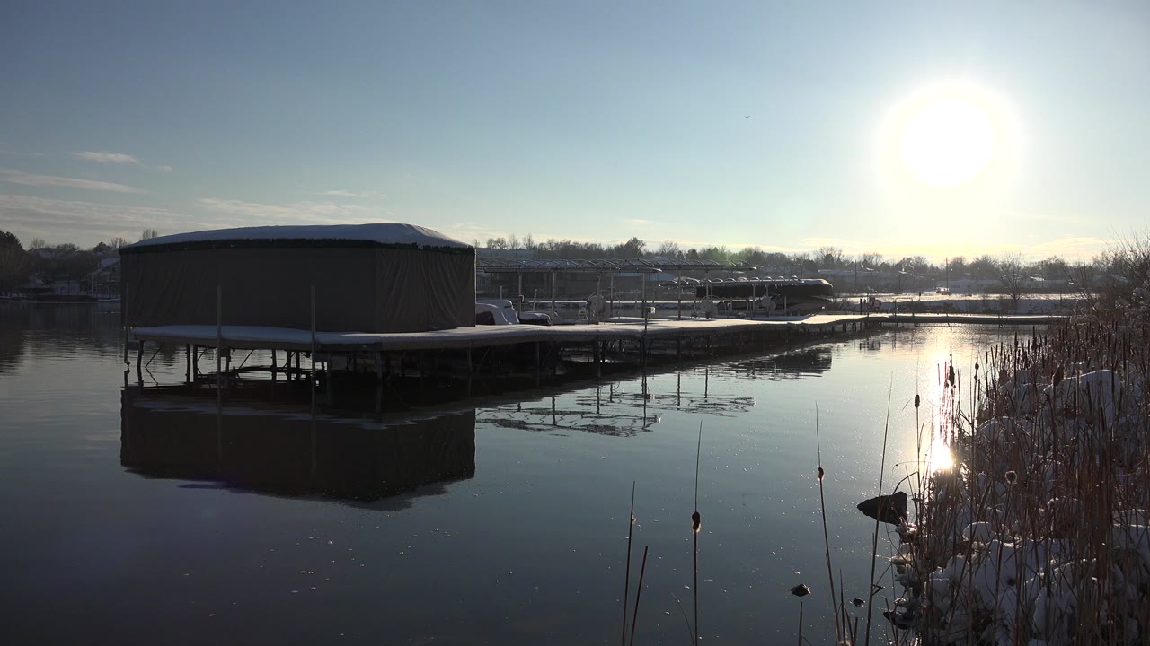 Covered boat dock remains centered over calm Westminster lake water, with cattail-like reeds in the foreground, bright sun high in frame, and no visible people.