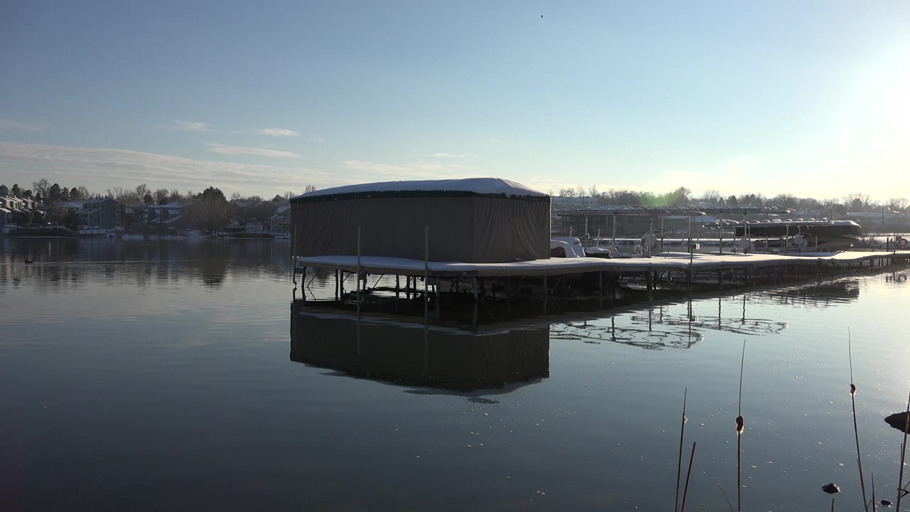 A covered dock sits across calm pond water in Westminster, Colorado, with its dark reflection below and a line of houses and trees beyond.