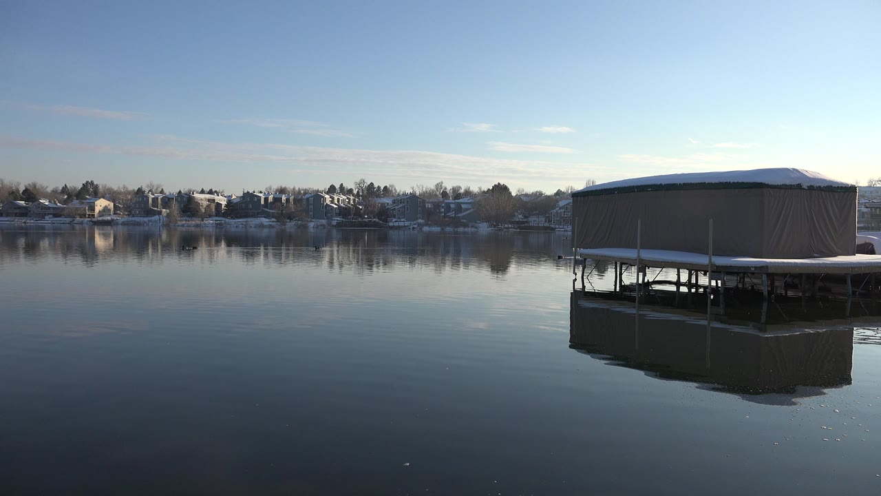 Westminster, Colorado lake view with the tan structure slightly right of center, its supports reflected in glassy water, distant snow-covered houses, and no visible people.