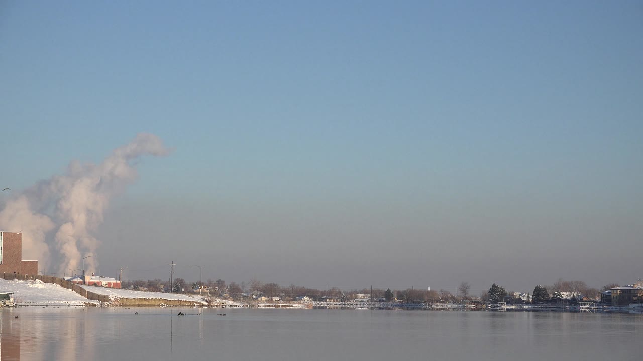 Westminster, Colorado shoreline with a brick building at left, a large pale plume rising behind the snowy bank, calm water below, and no visible people.