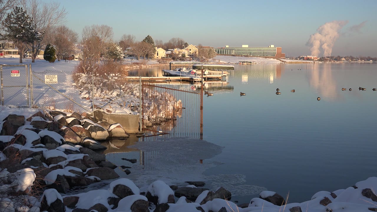 At 0:00 in Westminster, Colorado, snow-covered rocks and reeds border a calm pond with ducks on the water, houses and docks across the shore, and no visible people.