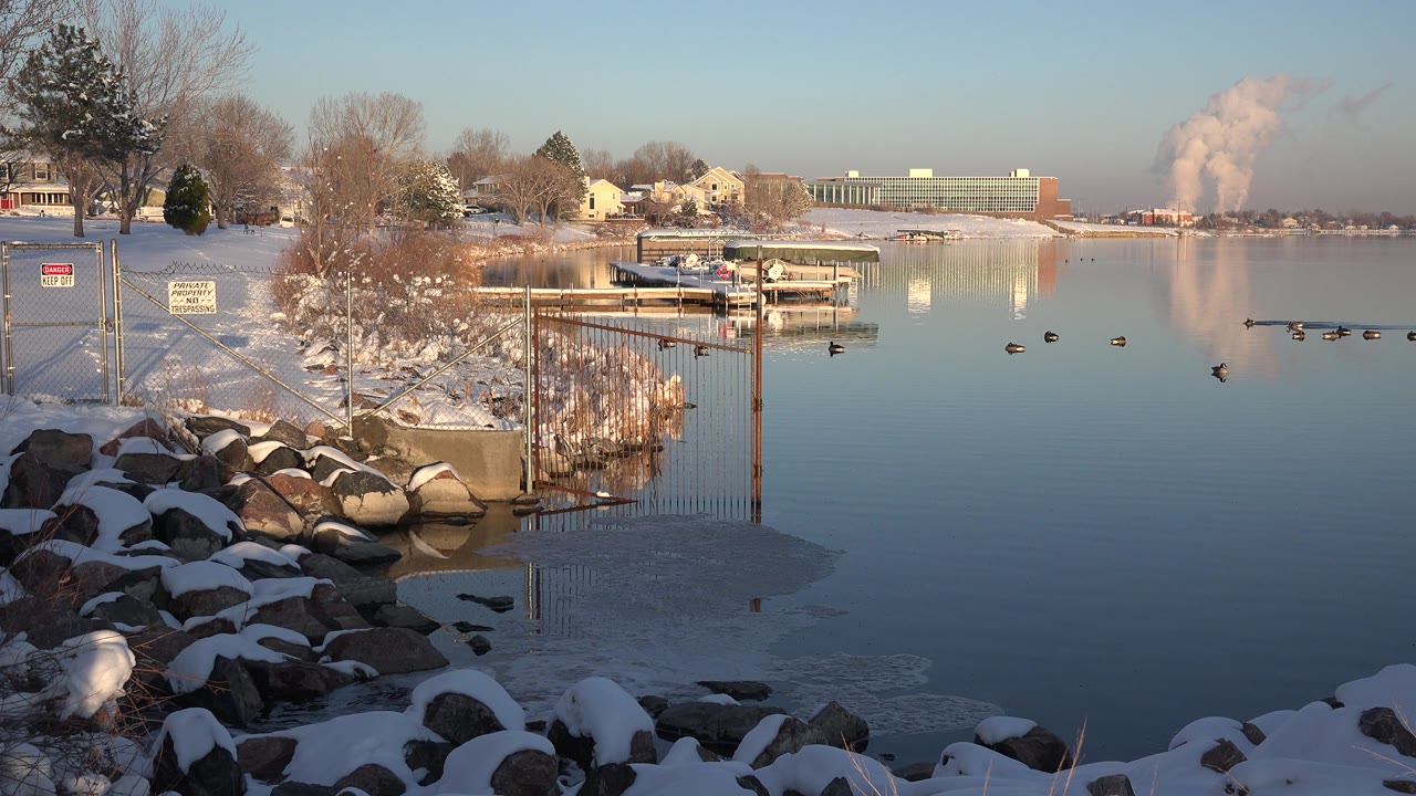 Opening frame of a Westminster, Colorado winter waterway with snow-covered shoreline rocks, ducks on still water, distant buildings, and no visible people.