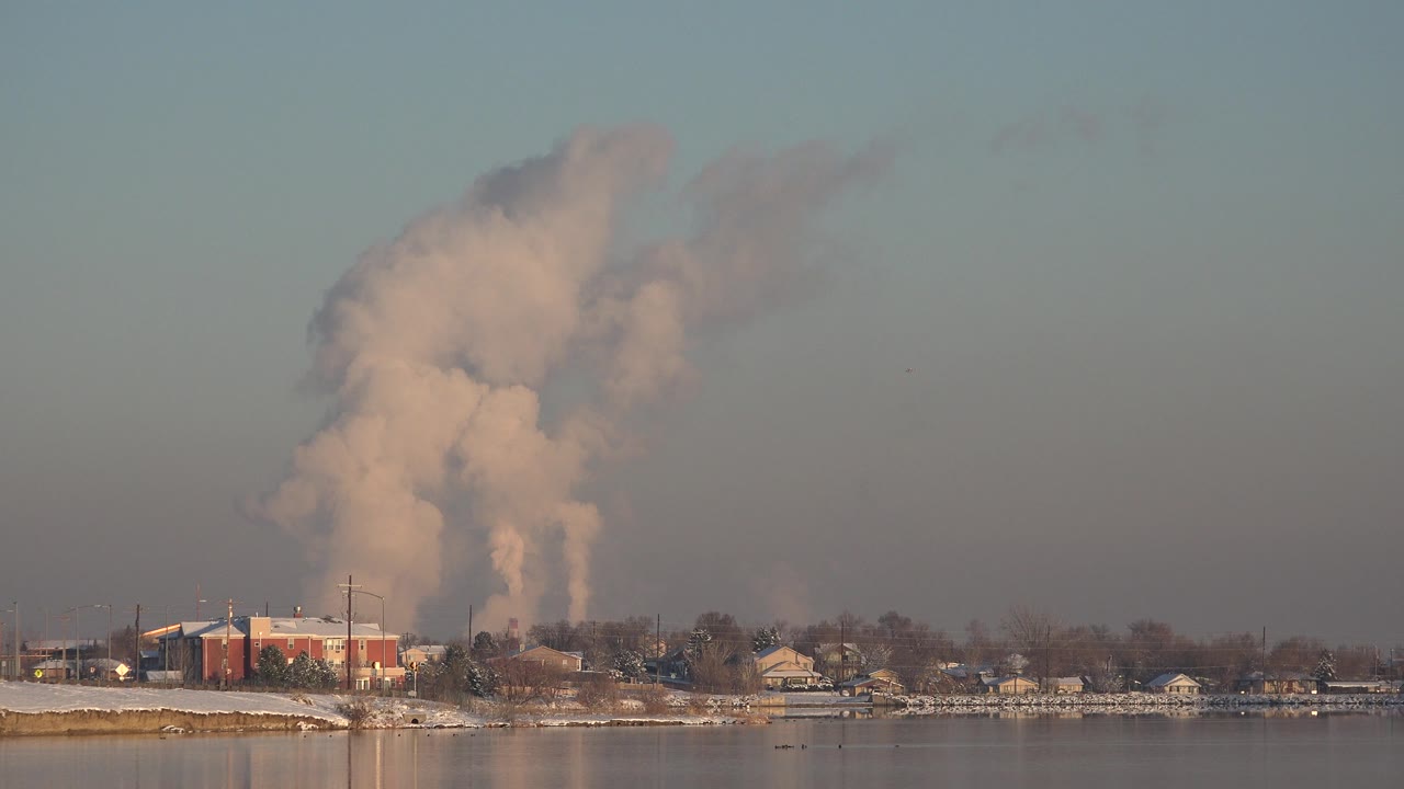 Smoke plume rising above a red industrial building across water.