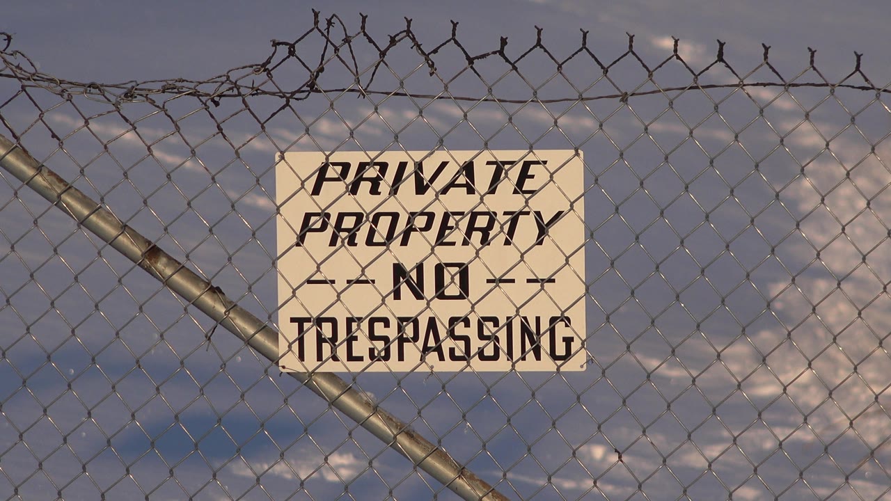 Weathered private property sign behind chain-link fencing and barbed wire.