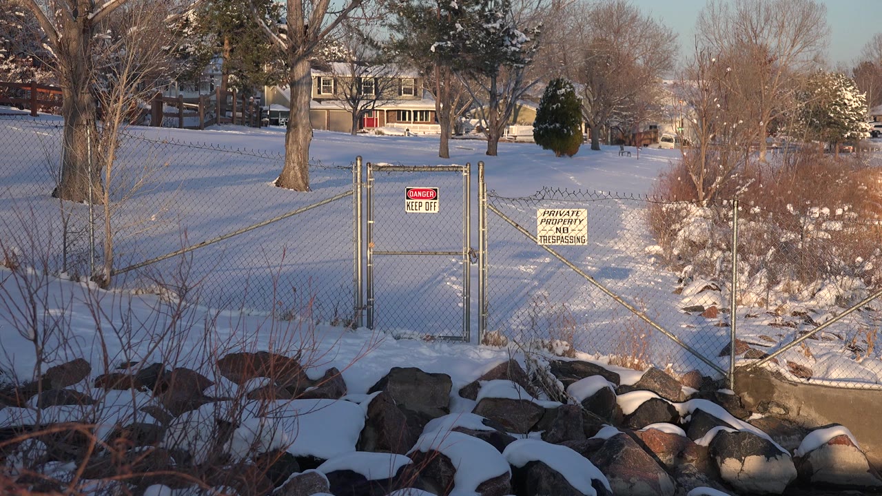 Snow-covered rocks and brush lead to a chain-link gate with warning signs beside a winter water edge in Westminster, Colorado, with houses and bare trees in the background and no visible people.