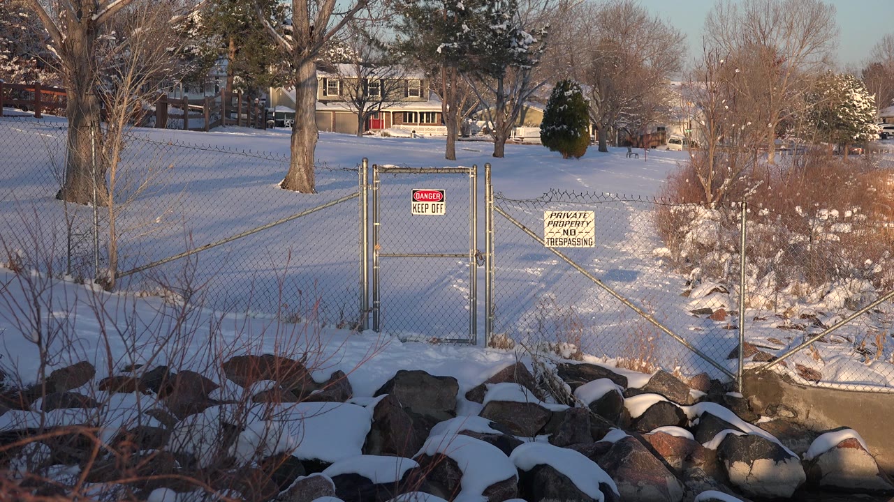 A slightly shifted view of the snowy Westminster shoreline centers the chain-link gate and posted signs, with rocks in front, shrubs to the right, homes beyond the trees, and no visible people.