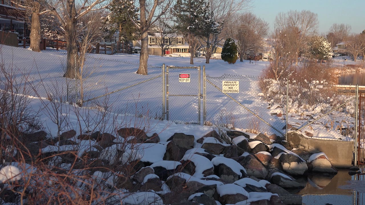 Fenced snowy shoreline beside calm water with rocks, brush, trees, houses, and posted signs.