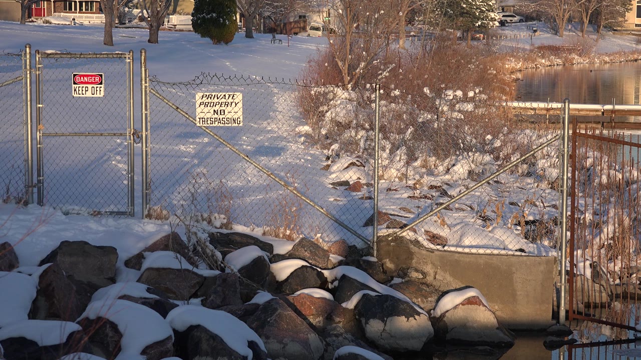 Snowy chain-link fence beside a calm lake with rocks in front.