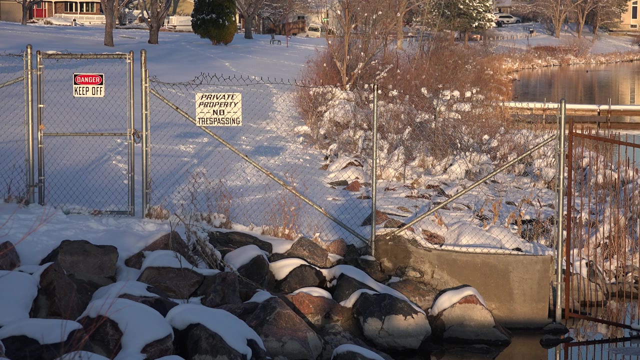 The second frame keeps the Westminster lakeside fence prominent, with snow on boulders, private-property signage, leafless trees, still water, and no visible people.