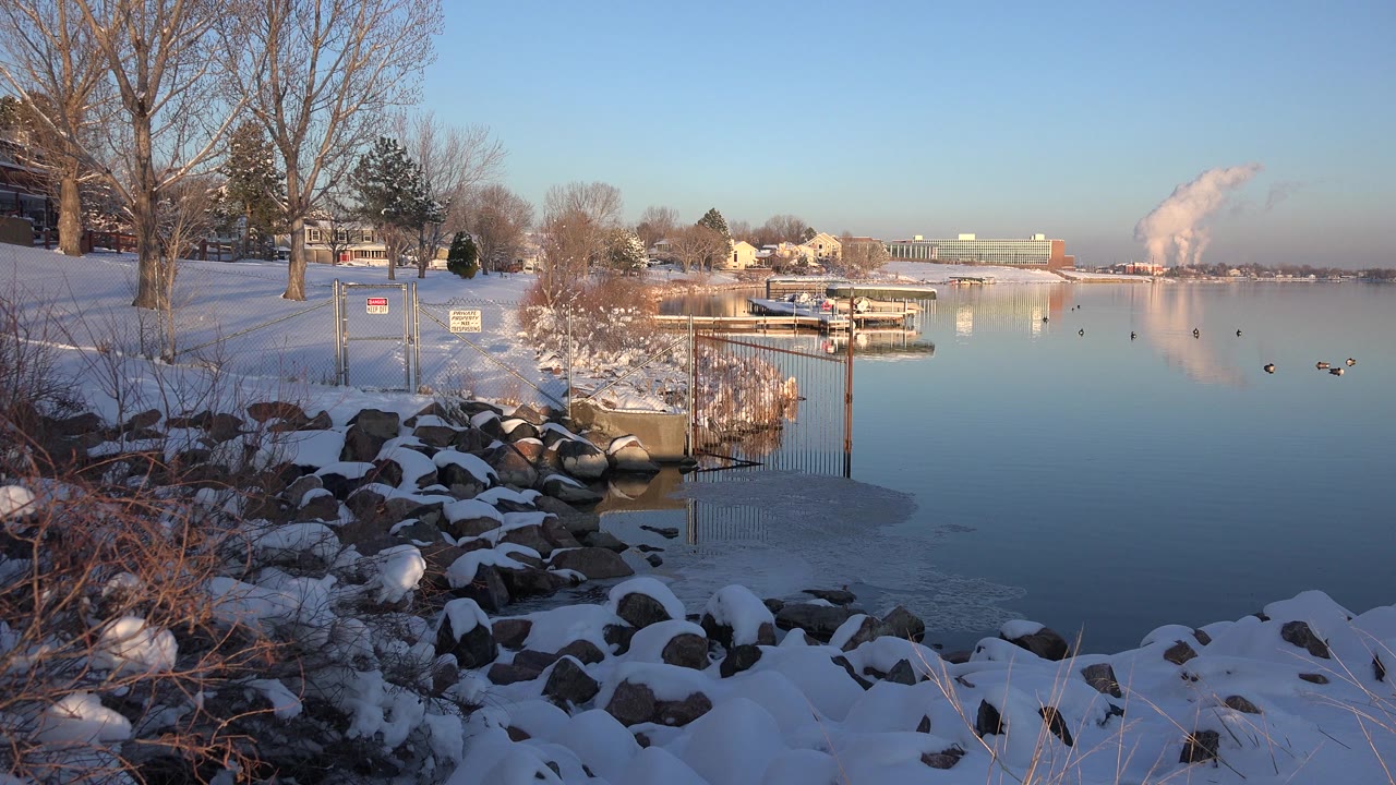 Westminster, Colorado lake view at the start of the clip, with snow-covered rocks in the foreground, bare trees at left, docks across the water, ducks on the lake, and no visible people.