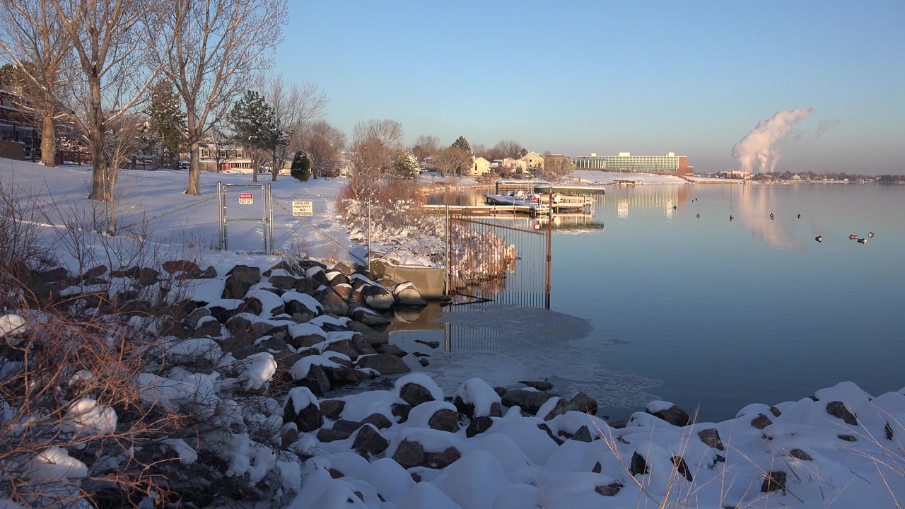 Westminster, Colorado winter shoreline with dark rocks under snow, brown brush at the water's edge, a docked lakeside structure, ducks on reflective water, and no visible people.