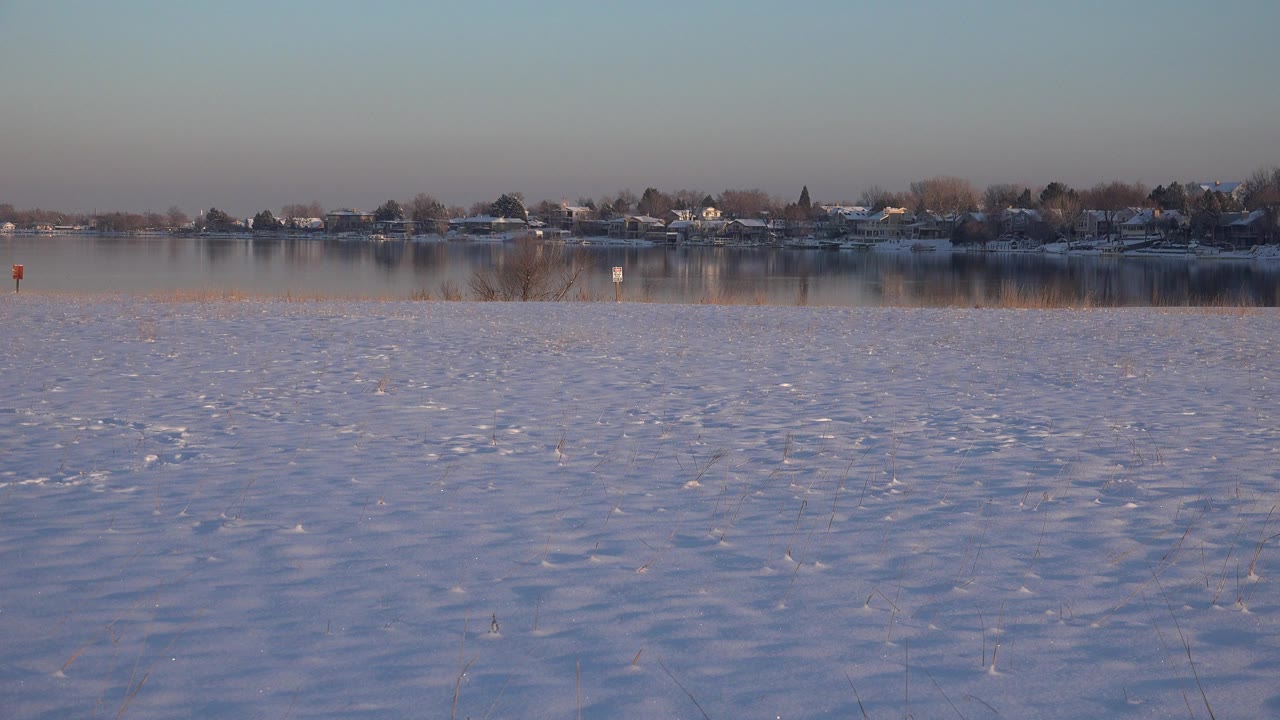 Wide Westminster, Colorado winter view at the start of the clip, with snow in the foreground, still water in the midground, distant buildings and trees, and no visible people.