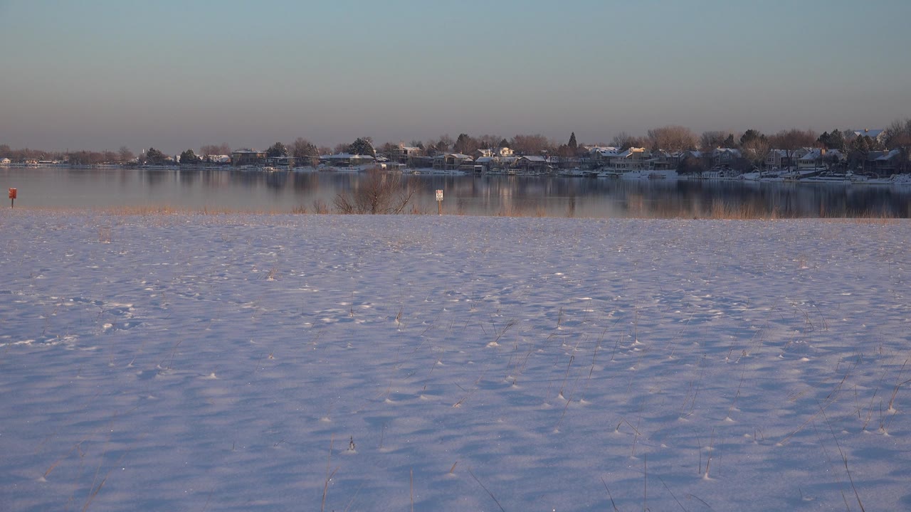 Second frame of the Westminster winter scene, with textured snow across the foreground, calm water beyond it, a tree-lined row of buildings, and no visible people.