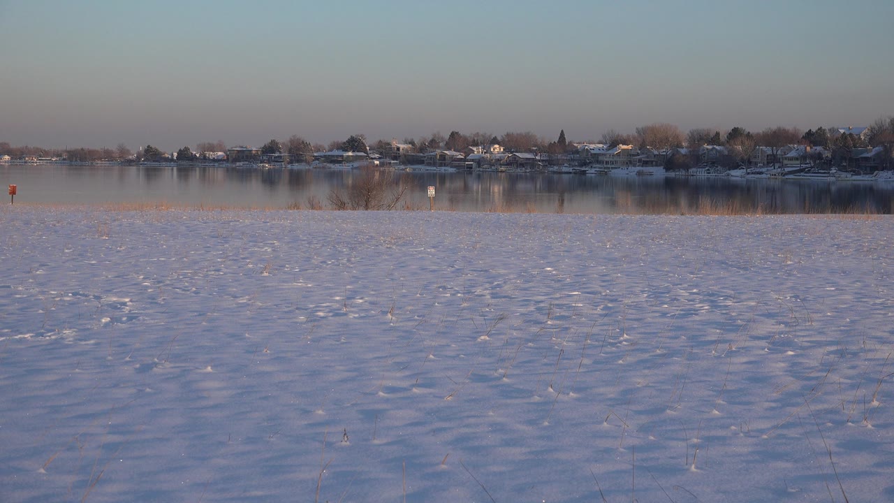 Snowy riverbank with water and distant houses across the shore.