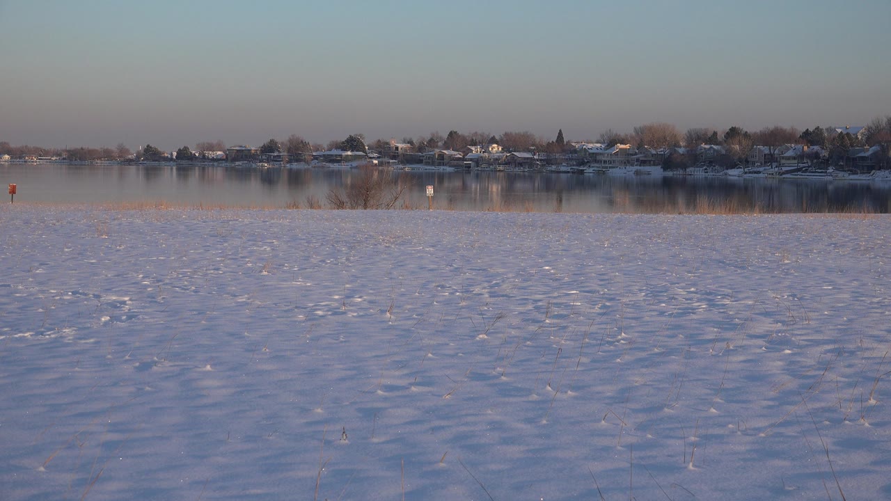 Snowy ground beside still water with distant homes and trees