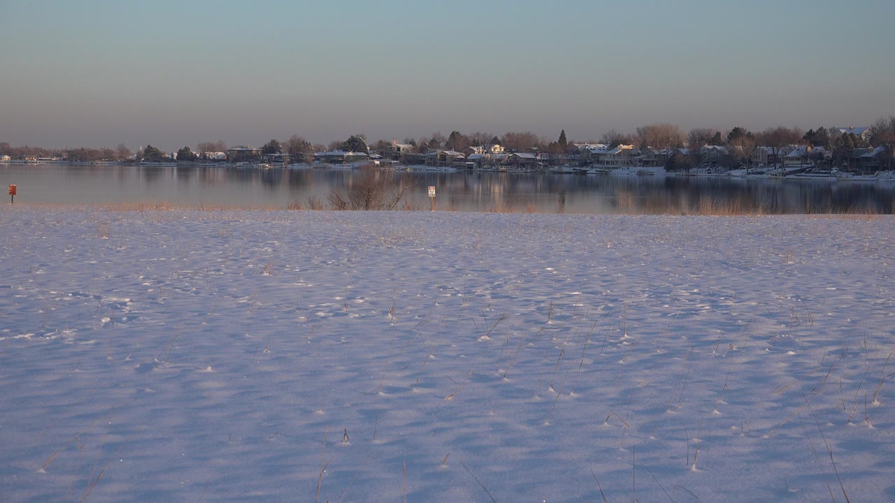 Snowy foreground fills the lower frame with calm water and a distant Westminster, Colorado residential shoreline beyond it, with no visible people.