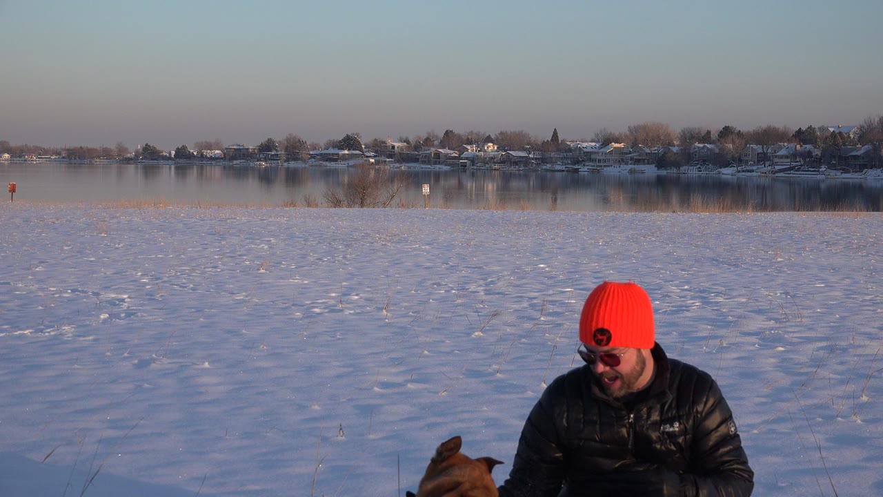 In Westminster, Colorado, a man in a bright red knit hat crouches near a partly visible brown dog on a snowy lakeside field with houses across the water.