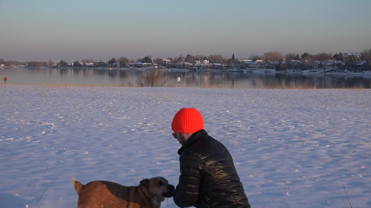Man in red hat turns toward a brown dog standing in the snow.