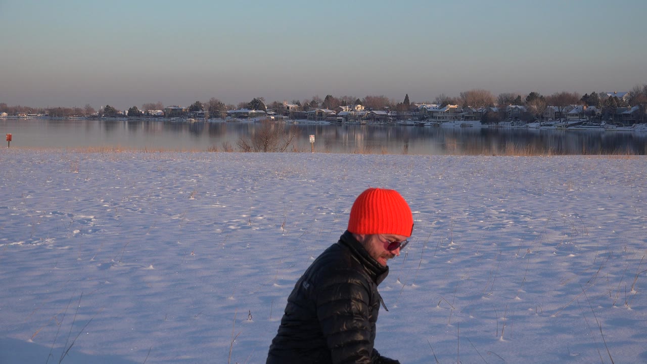 Man in red knit hat walking through snow beside a lake.