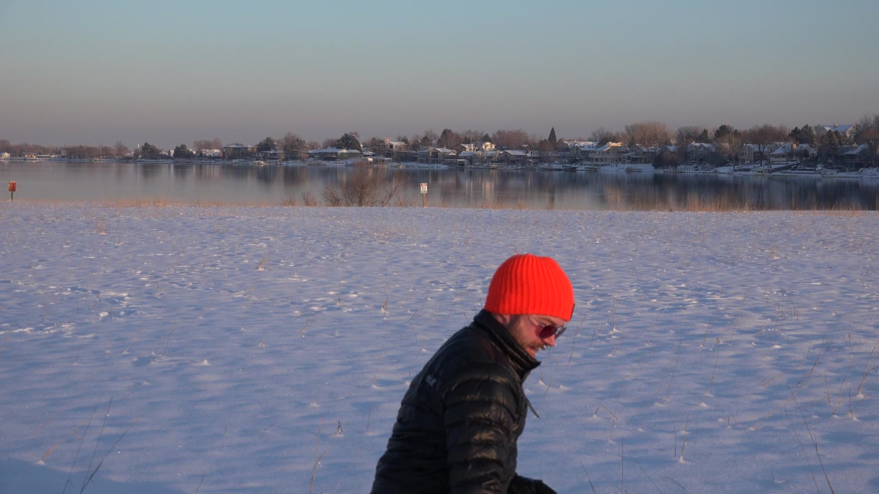 The man in the red knit hat is seen in profile against a wide snowfield, with the lake and distant homes behind him in Westminster, Colorado.