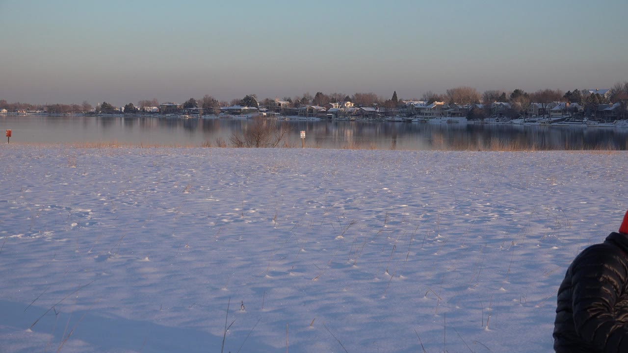 Snowy field beside a frozen lake with a person partly entering the lower frame.
