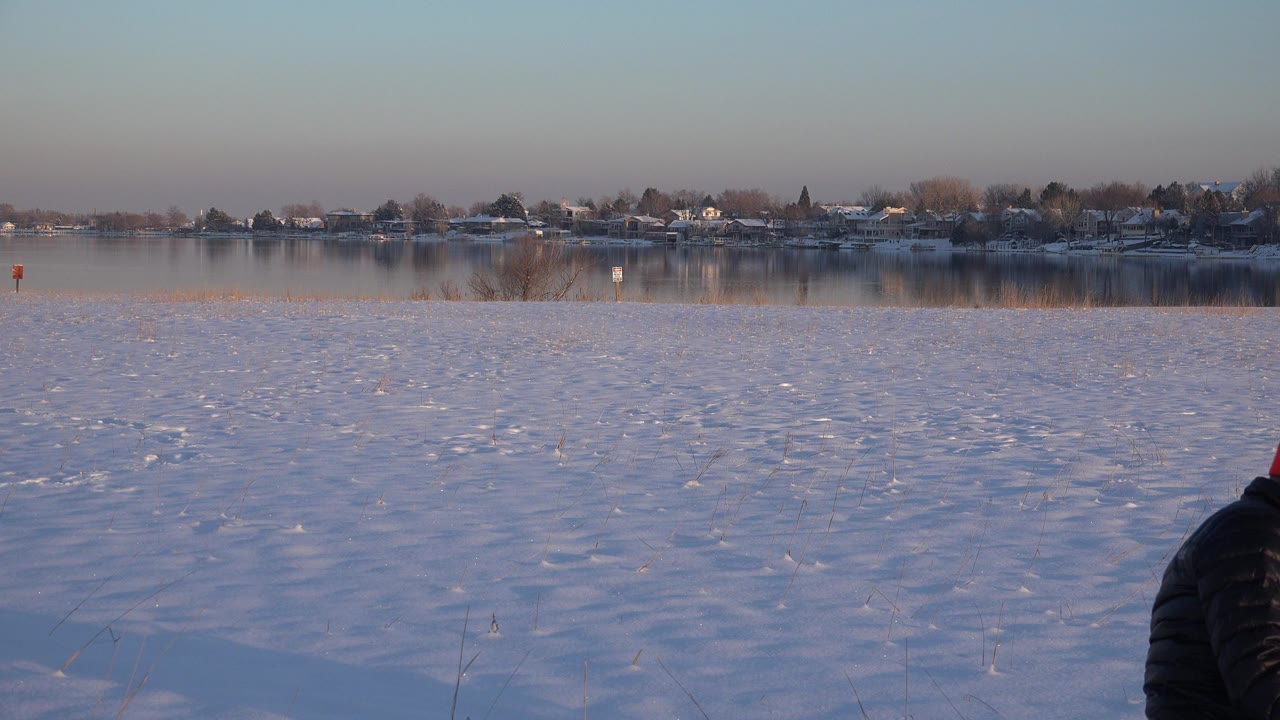 A partially visible person in a dark winter jacket bends near the bottom of the Westminster, Colorado frame, with snow, frozen water, reeds, and distant homes behind.