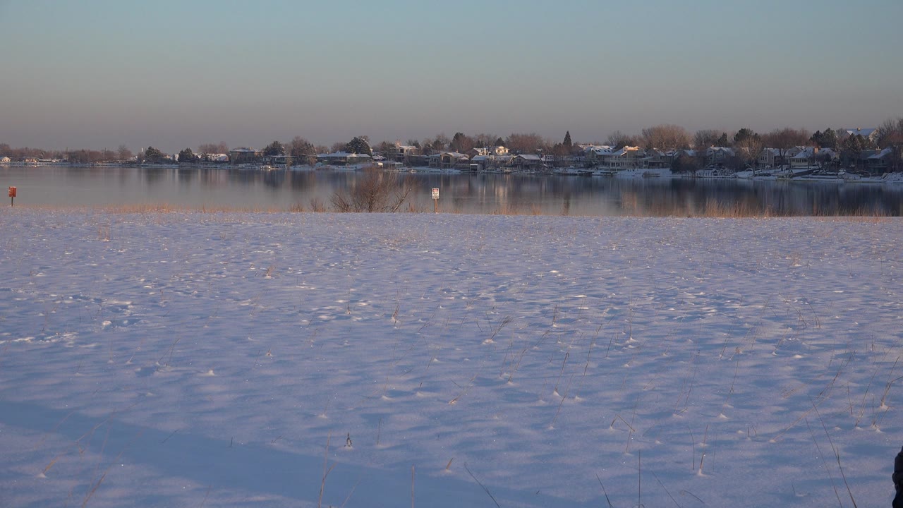 Wide view of snow-covered ground beside calm water in Westminster, Colorado, with distant homes and trees across the horizon and no visible people.