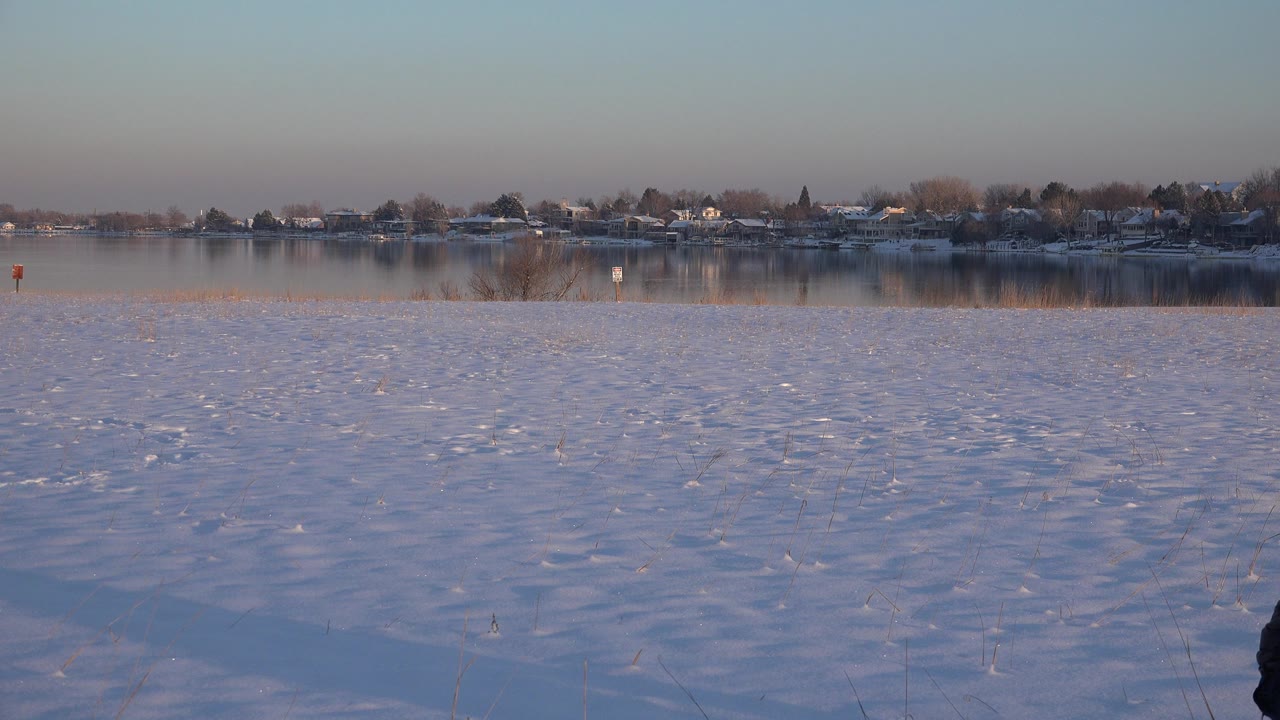 Snowy lakeside view in Westminster, Colorado, with distant homes across the water and part of a person appearing at the lower right edge.