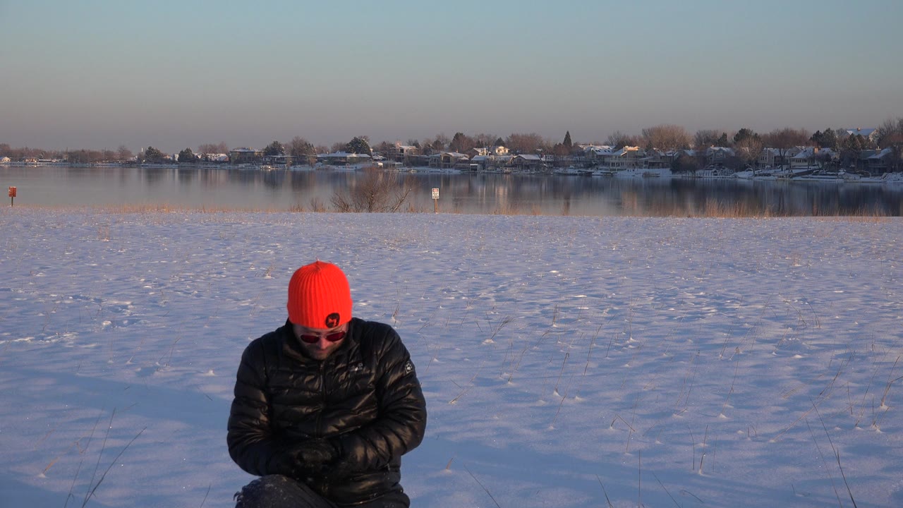 A man in a red knit hat and black jacket kneels low in snowy Westminster, Colorado, with a distant shoreline and homes across the background.