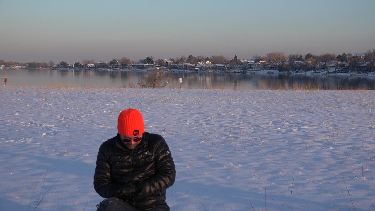 The man stays crouched in the snow in Westminster, Colorado, his red hat bright against the blue-white field and distant lakeside homes.