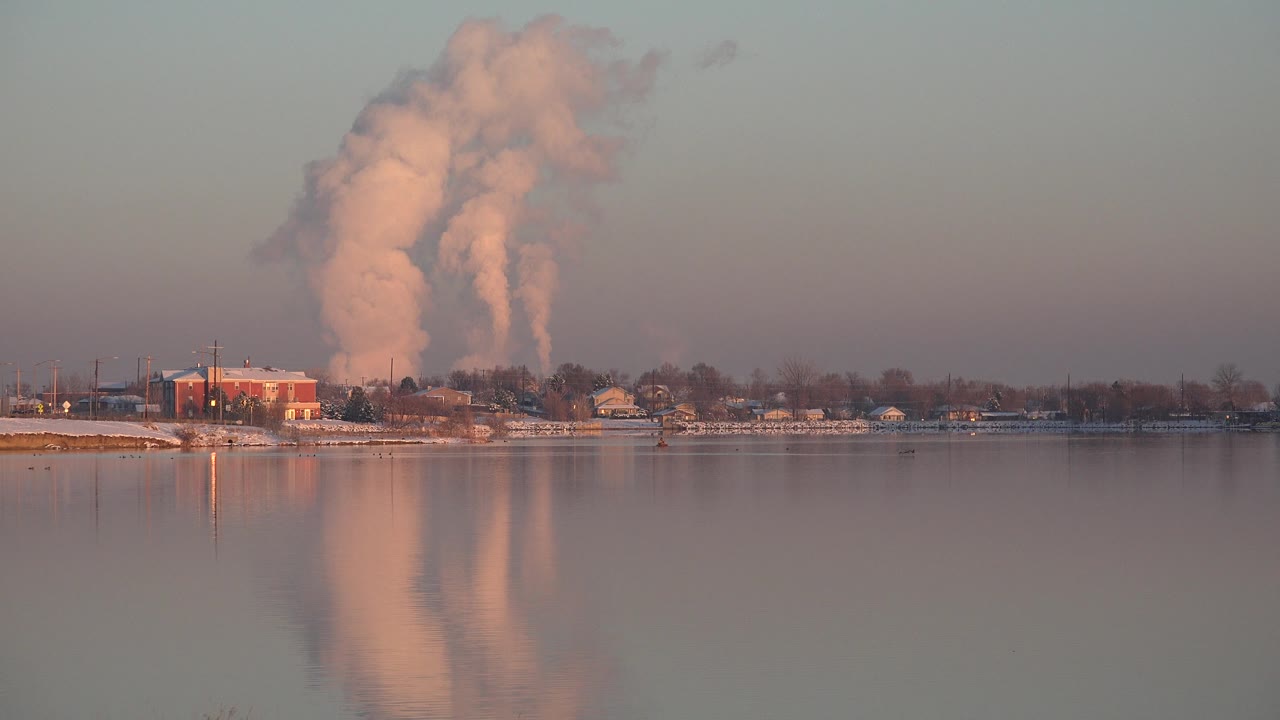 Wide view in Westminster, Colorado showing a large smoke plume rising behind low buildings and leafless trees, reflected in calm water, with no visible people.