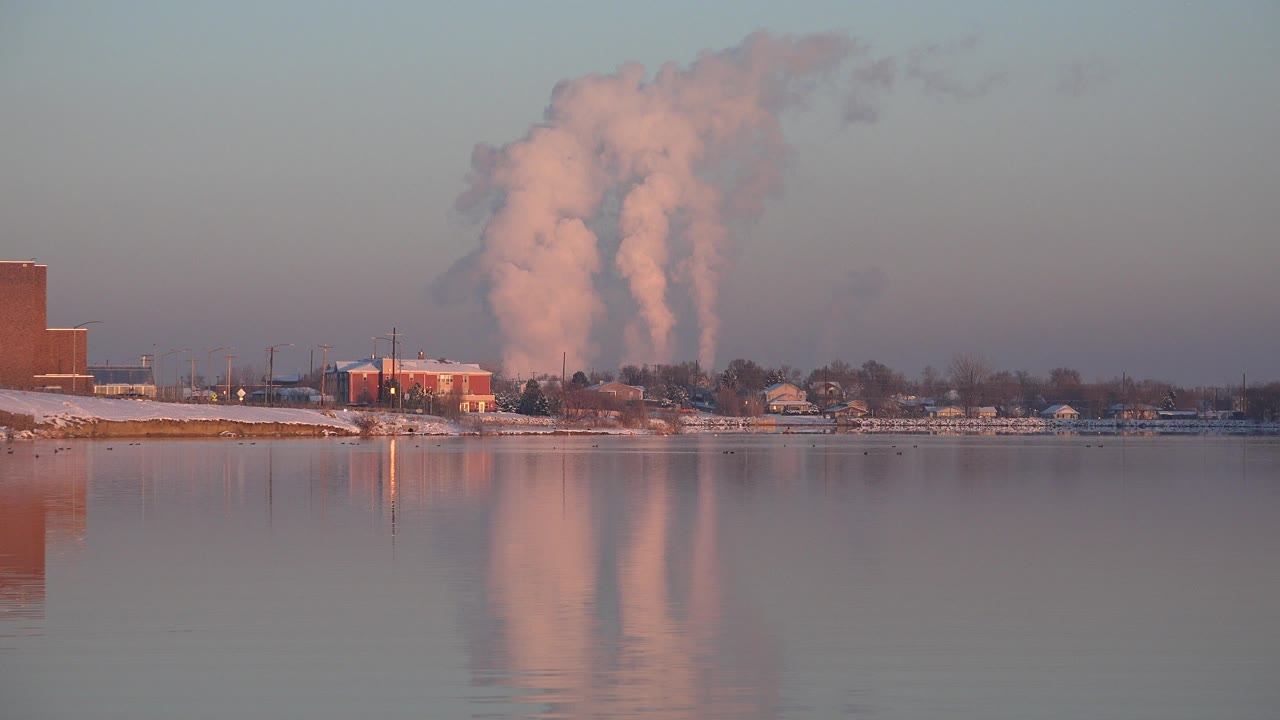 Opening frame of a large white smoke plume behind the Westminster, Colorado shoreline, with still water reflecting the pale cloud and no visible people.