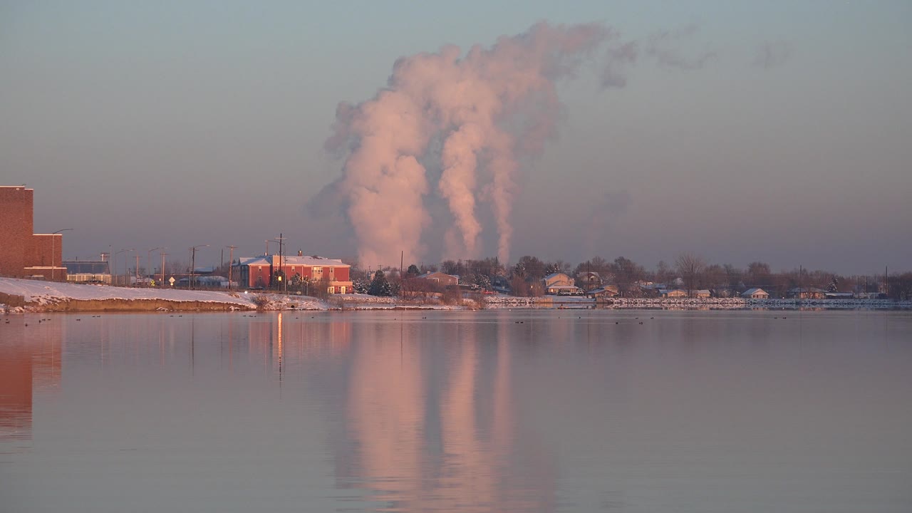 A dense white smoke column rises higher behind low buildings in Westminster, Colorado, with the plume reflected in the calm foreground water and no visible people.