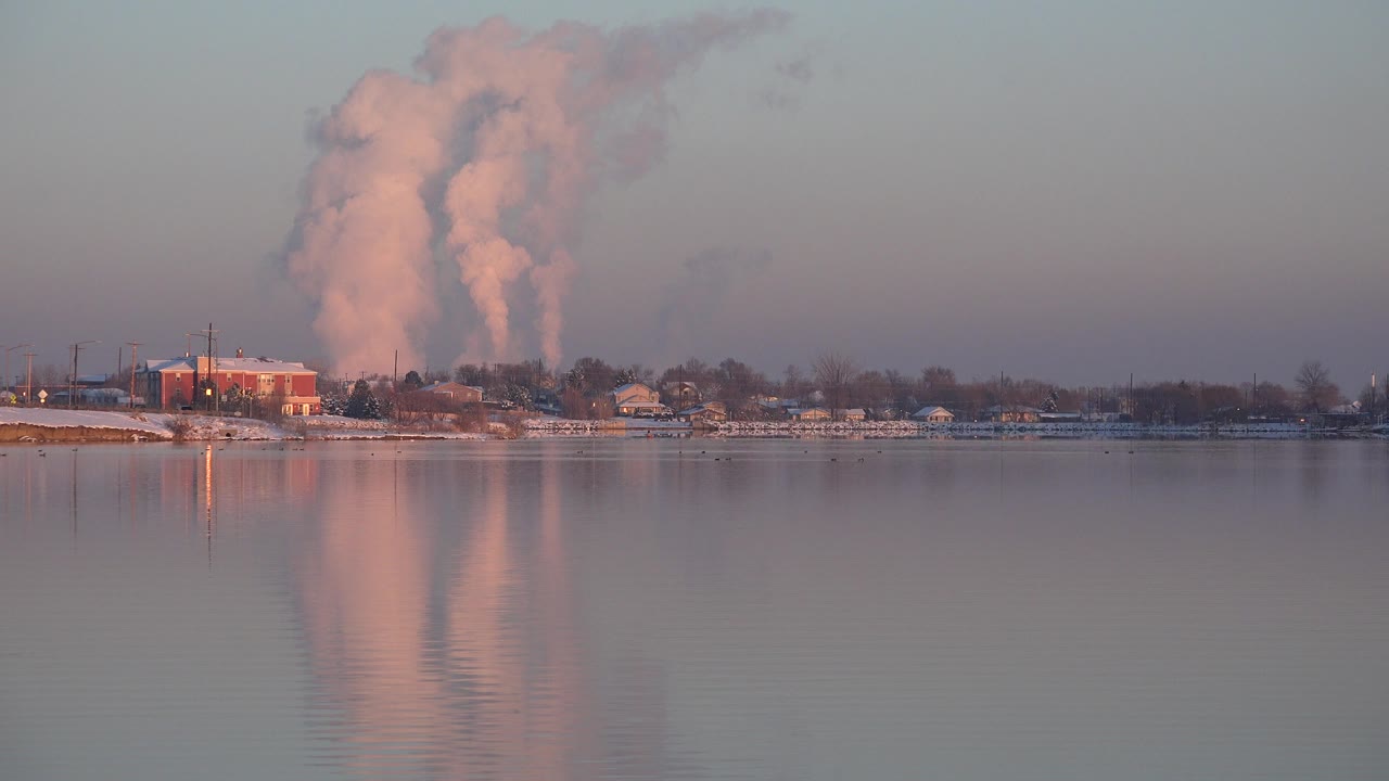 Opening frame of a Westminster, Colorado waterfront with calm reflective water, shoreline buildings, leafless trees, snow patches, and a pink-lit distant plume or cloud, with no visible people.