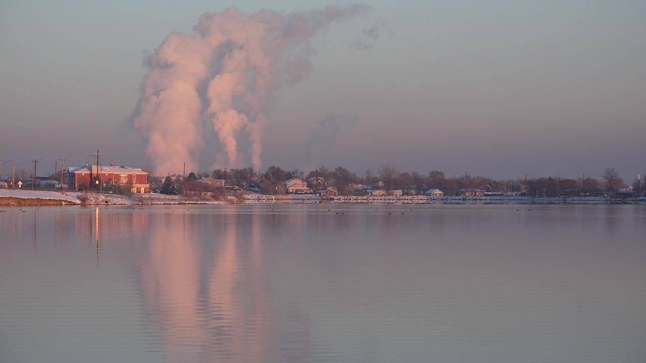 Calm water and low shoreline buildings with a large vertical plume or cloud in the distance.