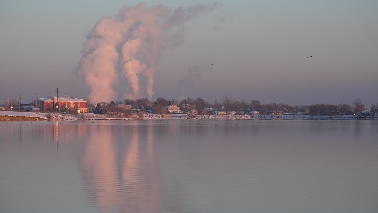 Factory fumes rise behind shoreline buildings across a lake.