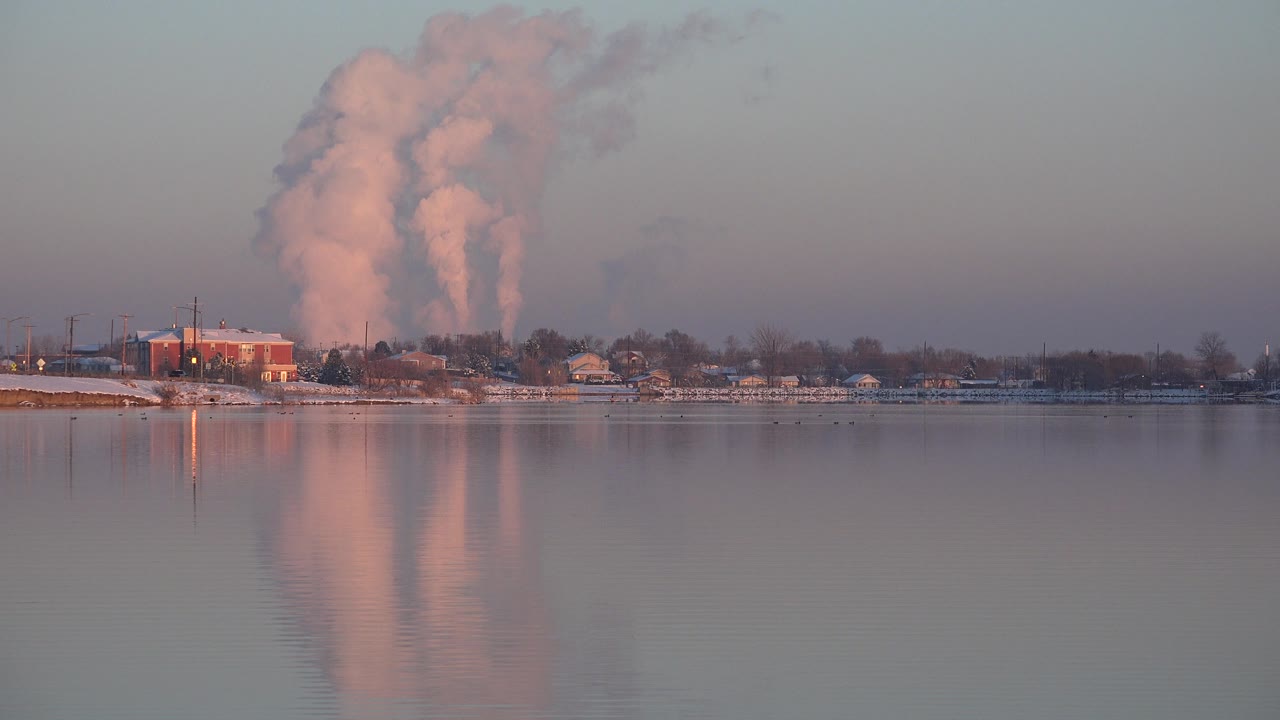 A wide Westminster, Colorado riverbank view shows calm water in front, low trees and buildings on shore, pink smoke plumes reflected in the water, and no visible people.