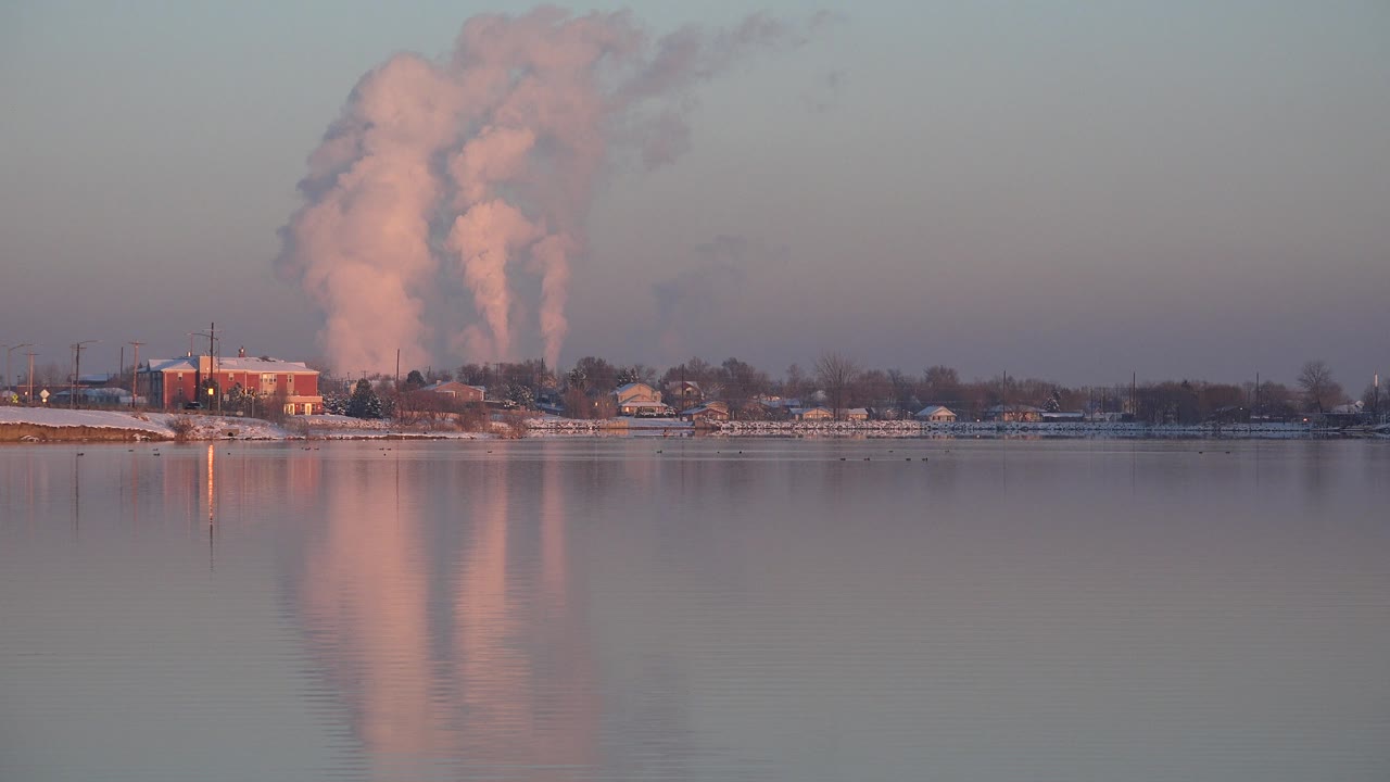 The Westminster river scene holds a broad water foreground while two warm-toned smoke columns rise behind shoreline trees and low structures, with no visible people.