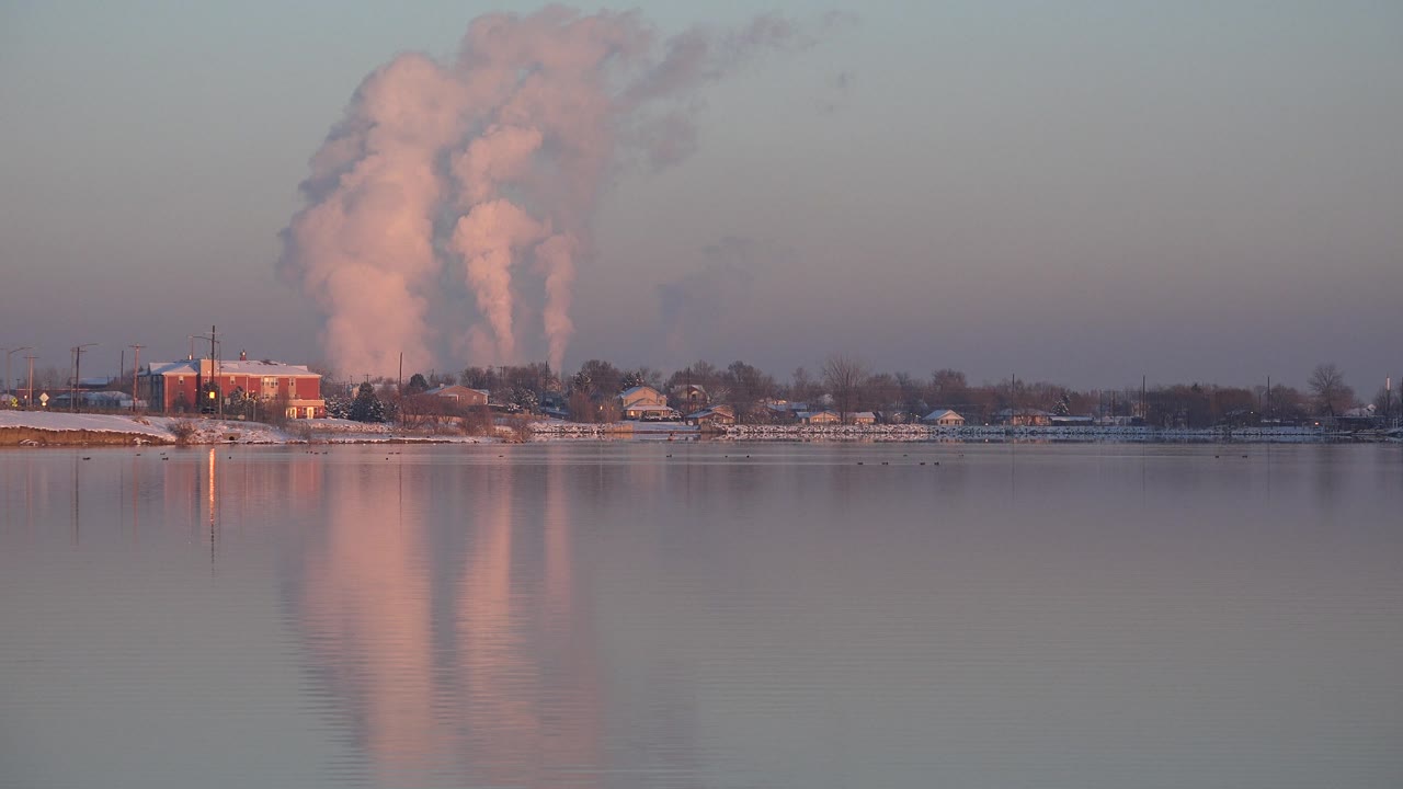 Sunset-lit smoke plumes rise above a distant industrial shoreline.