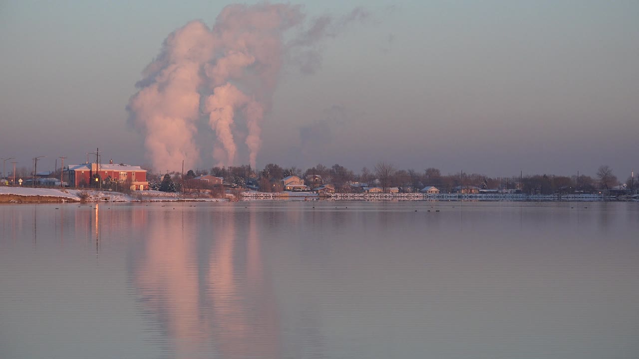 White smoke plume rising behind shoreline buildings across calm water.