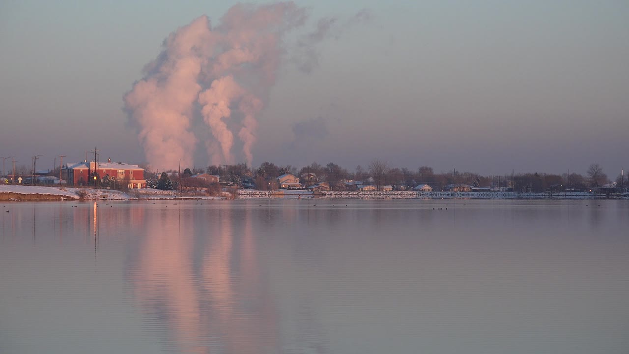 At 0:01, the Westminster, Colorado shoreline remains small across the frame while the smoke plume and its pink reflection become the dominant visual elements, with no visible people.