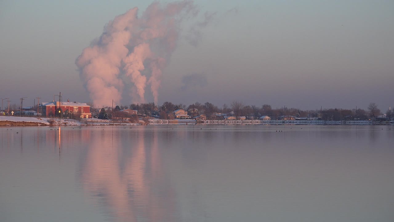 Distant shoreline buildings and a pink vapor plume reflected on still water.