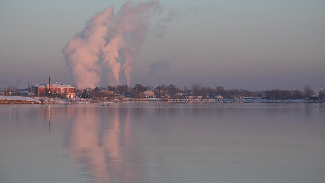 At 0:01 in Westminster, Colorado, the plume remains high above the shoreline while its pink reflection stretches down the calm water, with no visible people.